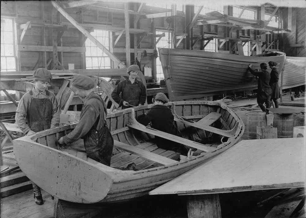 Photographie en noir et blanc – Intérieur d’un grand bâtiment en bois où des femmes en vêtements de travail, en salopettes, effectuent divers travaux pour la construction de bateaux en bois.